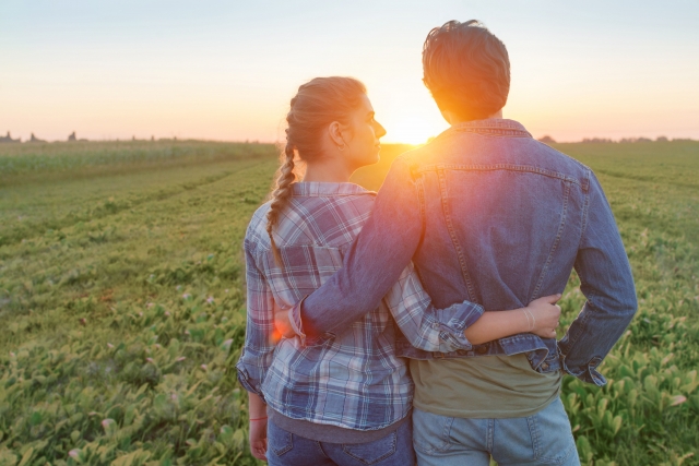 Ｇジャンを着た男性とジーンズを履いた女性が夕日を眺めながら肩を寄せ合う写真。